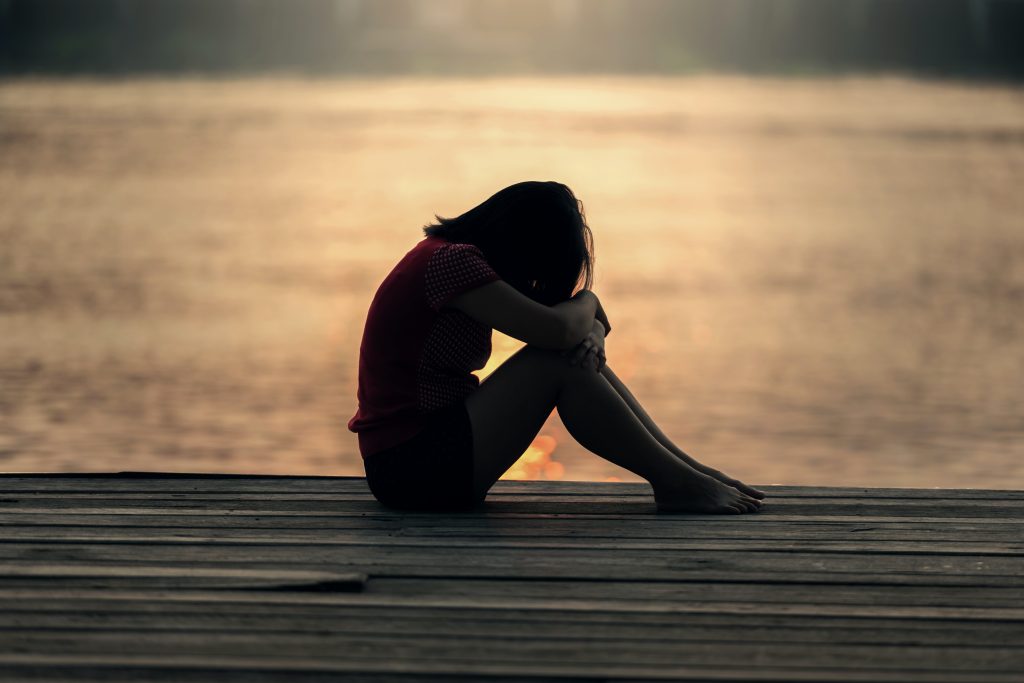 A despondent girl sits hugging her knees by the sea during sunset.