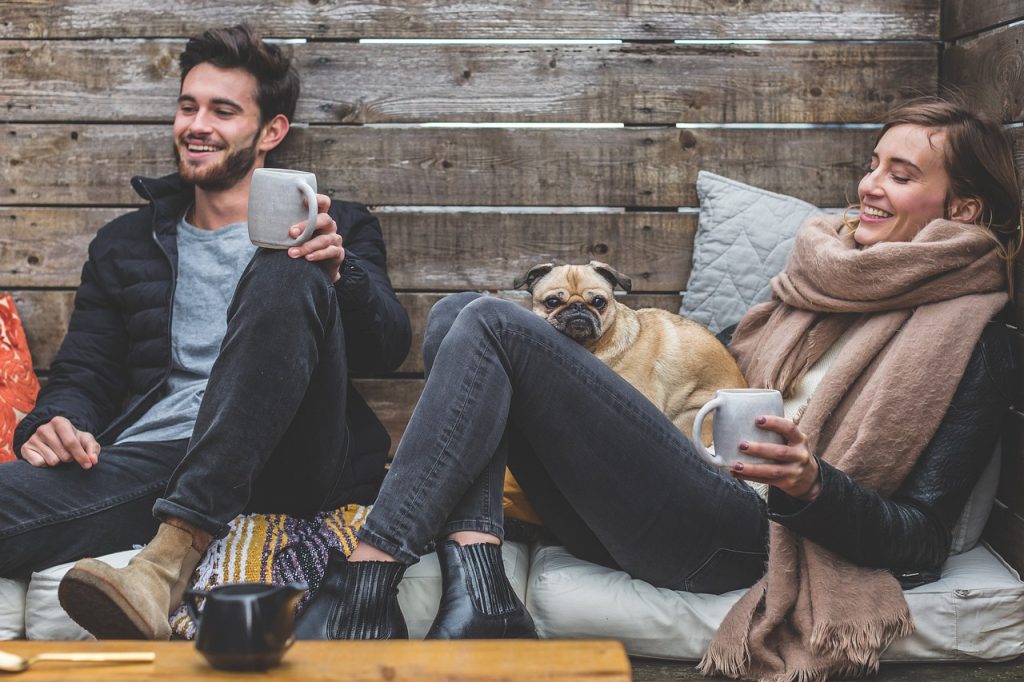 A man and a woman holding a dog sit on the sofa, enjoying coffee and laughing happily—they are friends.