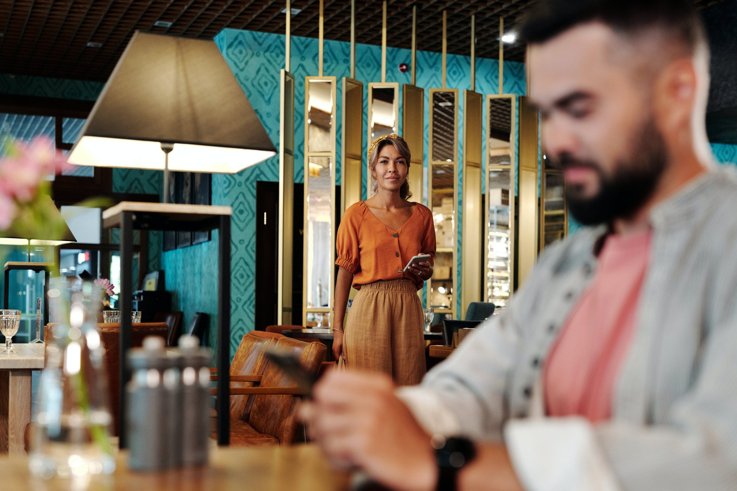 A man is sitting in the store waiting for someone with his mobile phone. A beautiful woman is holding her mobile phone and walking towards the boy with a smile，it's the first-date for them.