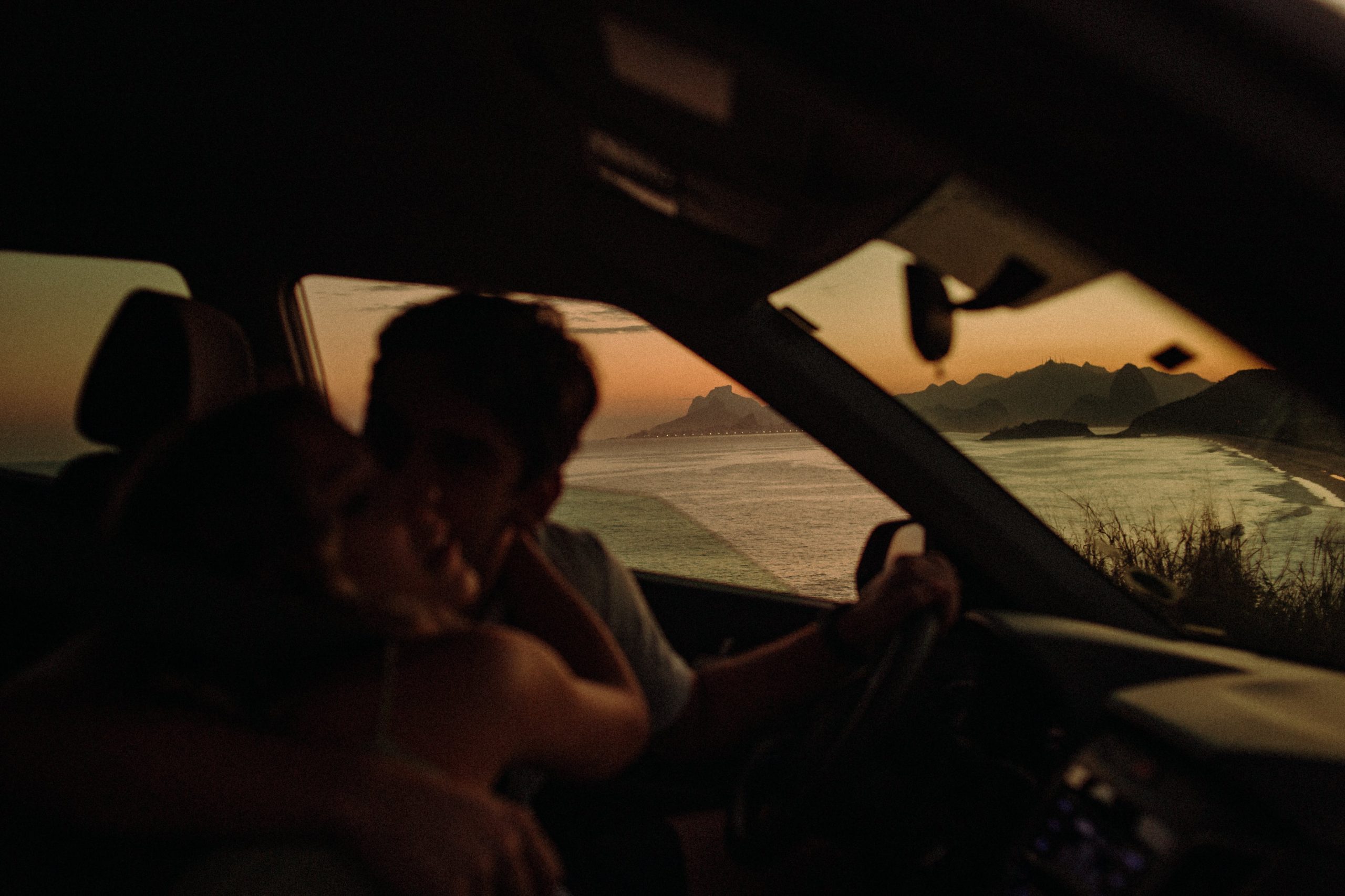 A couple sitting in a car, kissing each other on the cheeks under the rays of the setting sun.
