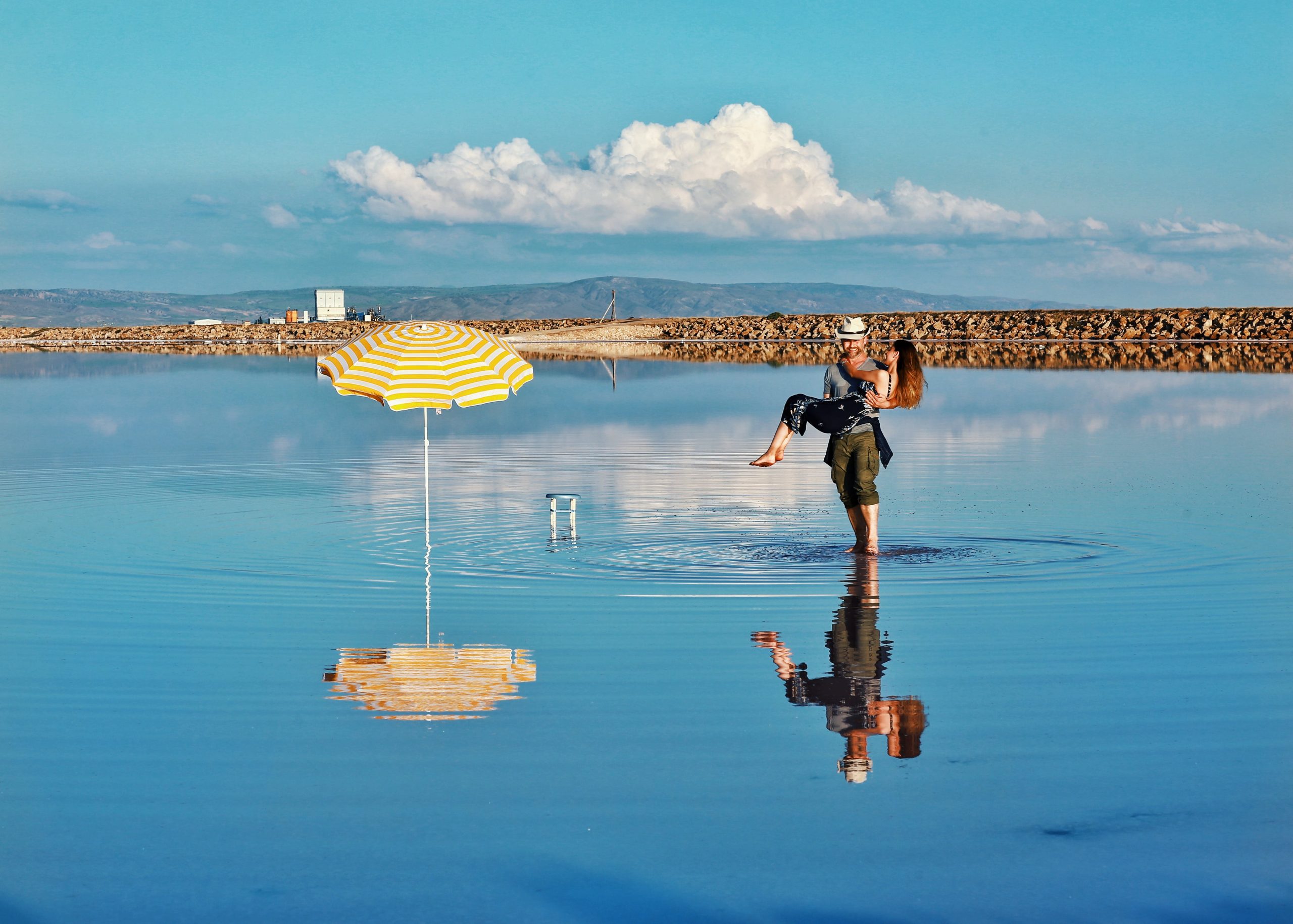 Beneath the azure sky and fluffy white clouds, the two embrace romantically while enjoying the picturesque scenery reflected in the tranquil lake.