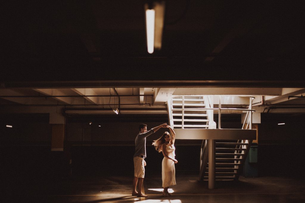 In a dark basement, a warm and bright beam of light shines through the darkness onto a couple dancing.