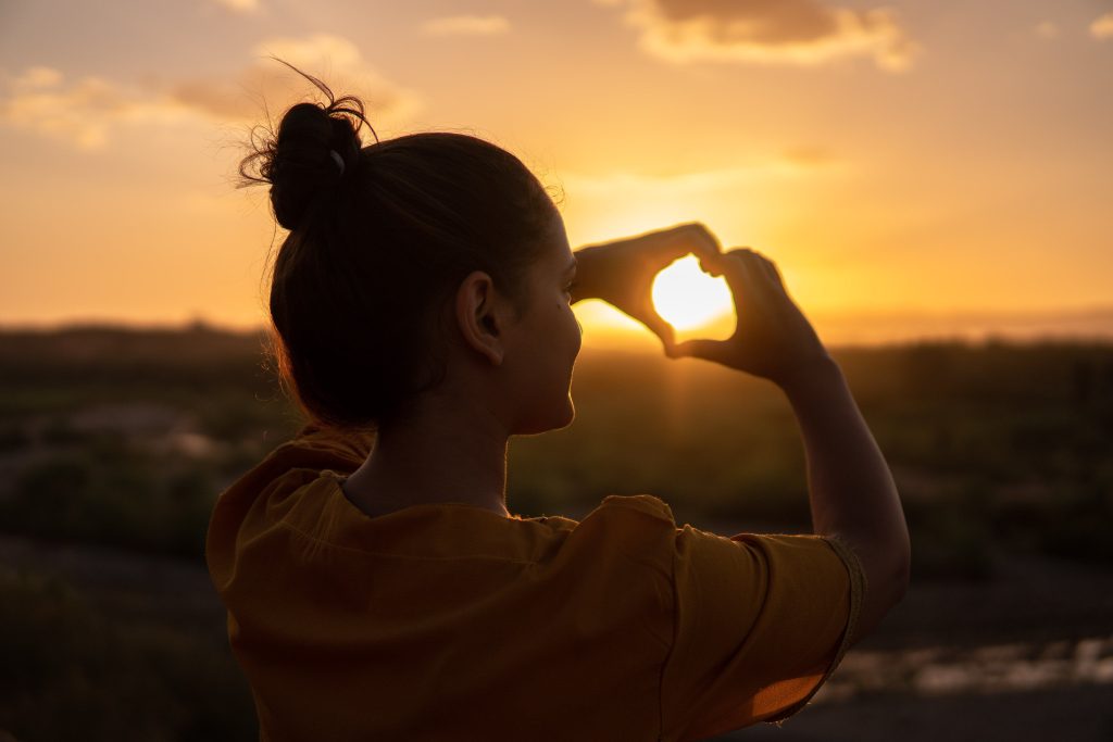 The girl makes a heart gesture under the sunlight.