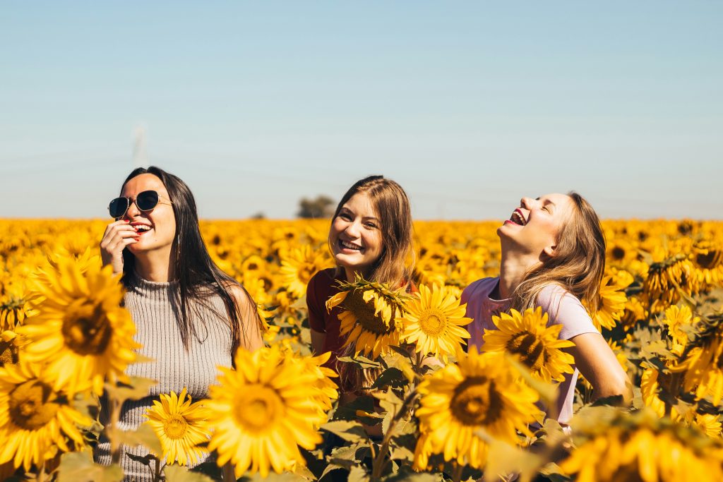 Three independent and confident women are smiling brilliantly among a field of sunflowers.