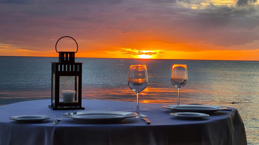 There is a dining table by the seaside, with the setting sun casting its rays on the transparent wine glasses, creating a romantic dinner setting.