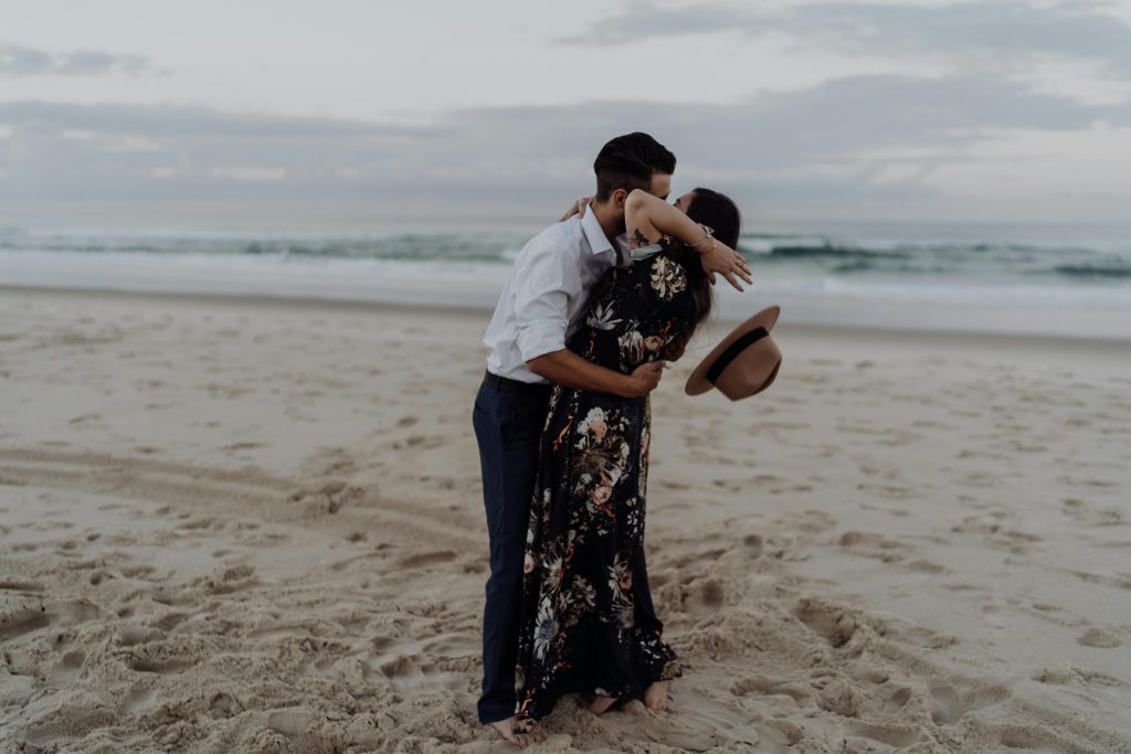 A couple is kissing by the seaside when a gust of wind blows, knocking off the woman's hat.