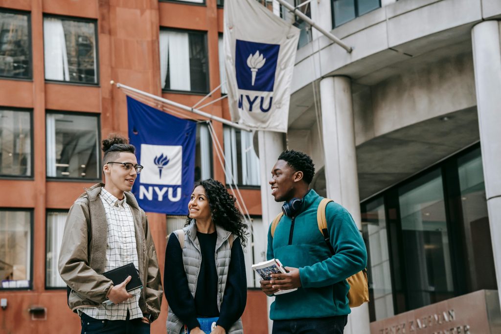 A female university student and two male university students chatting by the roadside, holding books.