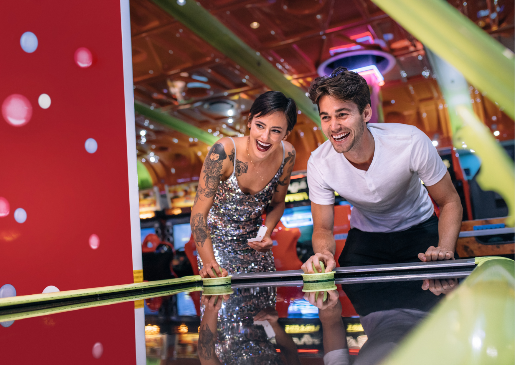 Happy couple playing air hockey in an arcade, laughing and enjoying a fun and flirty date night.