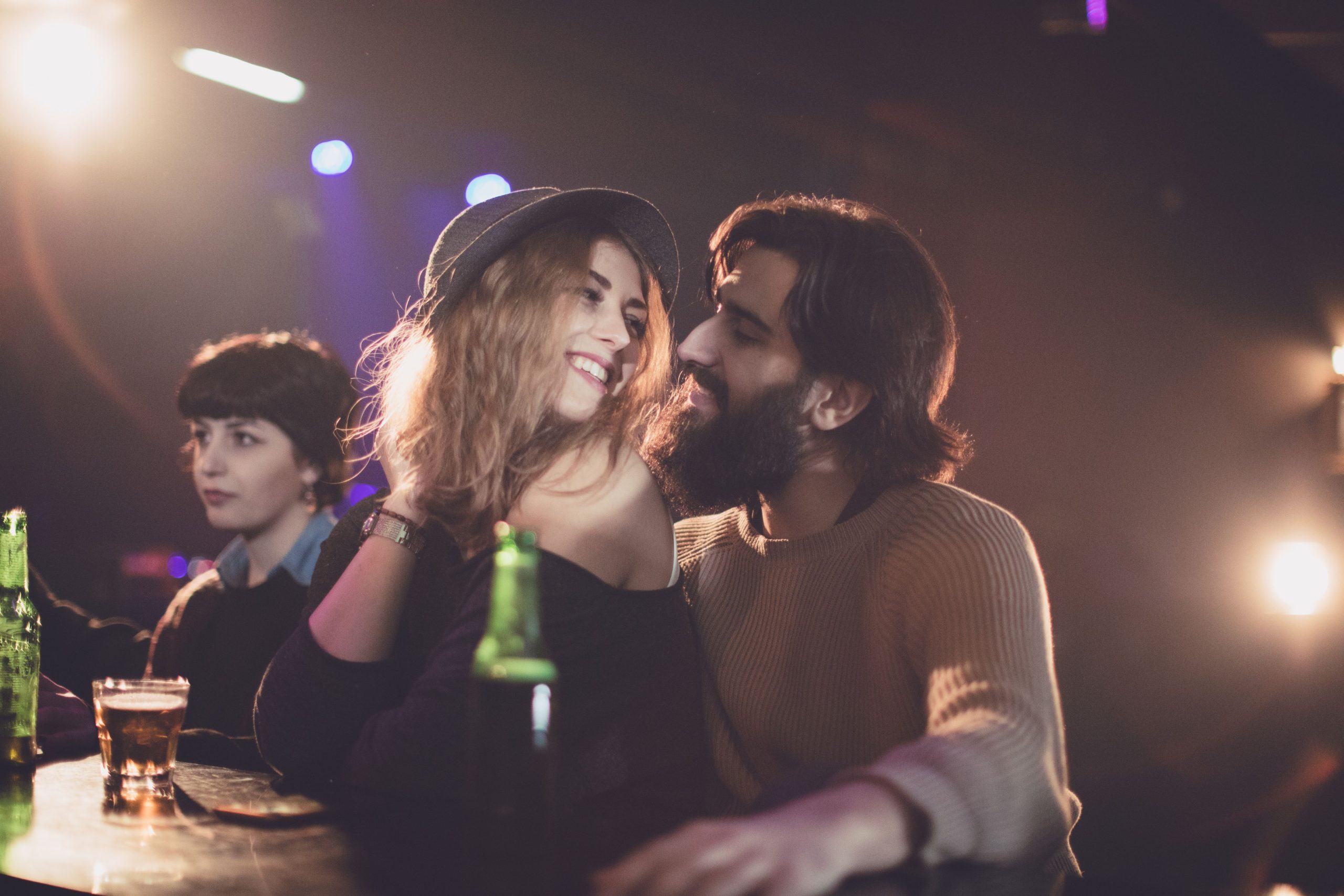 A flirty couple laughing at a bar, embodying the tension between casual and committed dating styles, with a third person observing in the background.